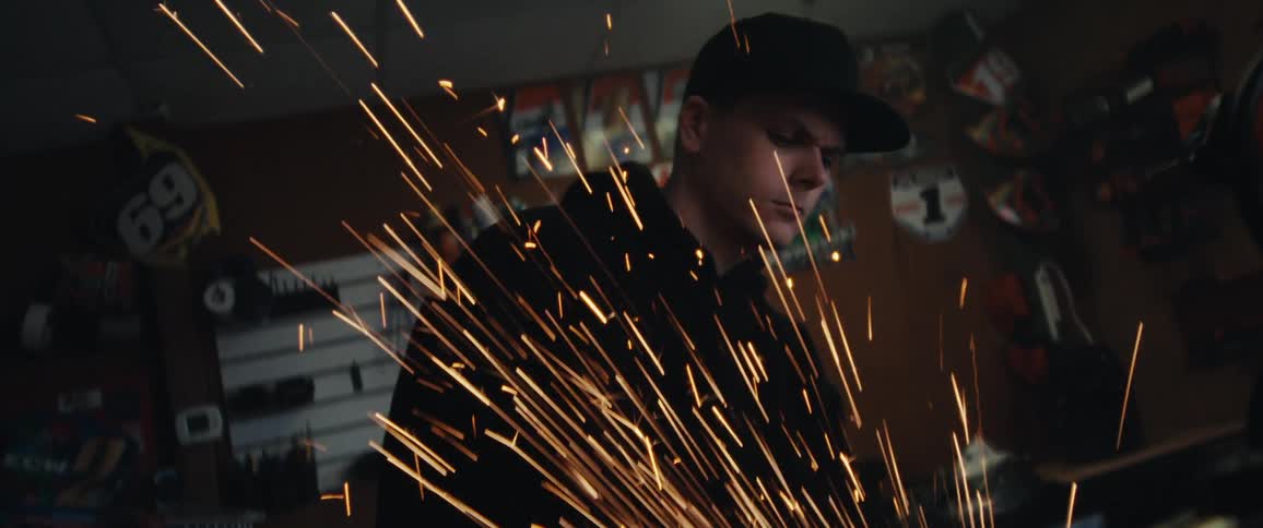 A mechanic grinds an orange dirt bike in a shop as the camera moves close to the shower of sparks