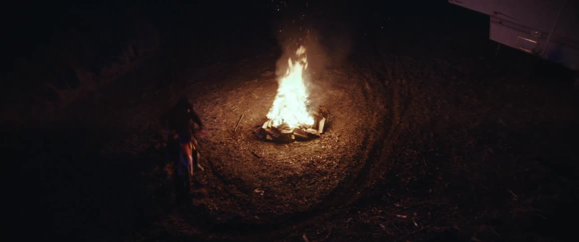 A motorbike intensely circles a roaring bonfire at night spraying dirt and etching tire tracks around the flames