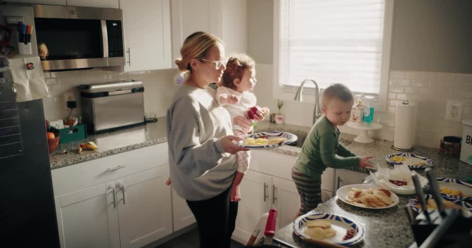 A mother holds a child and serves food on a plate in a kitchen while another child stands by the sink with plates of food on the counter