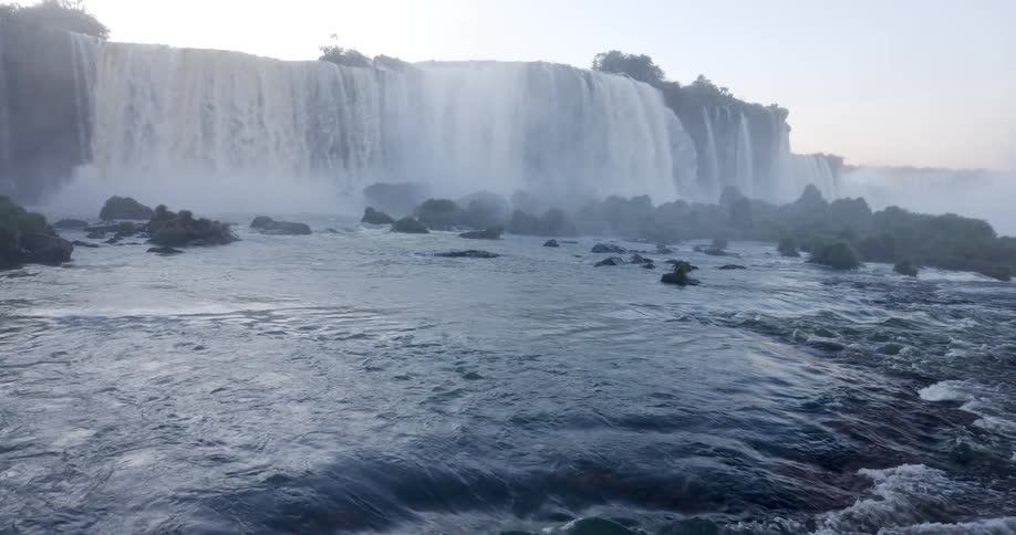 Water powerfully flows over a rocky waterfall generating mist in a natural outdoor scene with cliffside vegetation visible