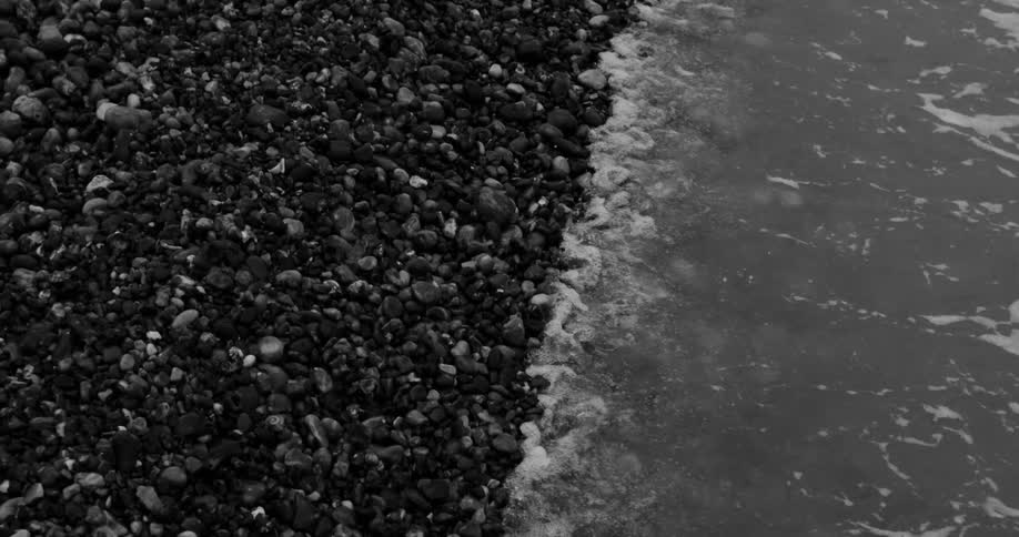 Aerial view of dark smooth pebbles meeting gentle sea waves at a shoreline
