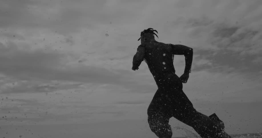 A person with dreadlocks is running through shallow water creating splashes against a cloudy sky backdrop
