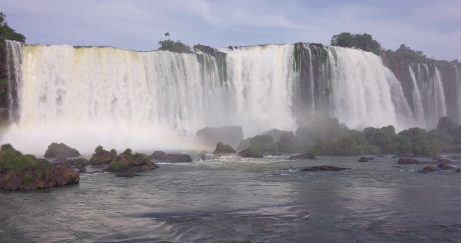 A wide multitiered waterfall cascades over a rocky cliff surrounded by lush forest with mist above the falls