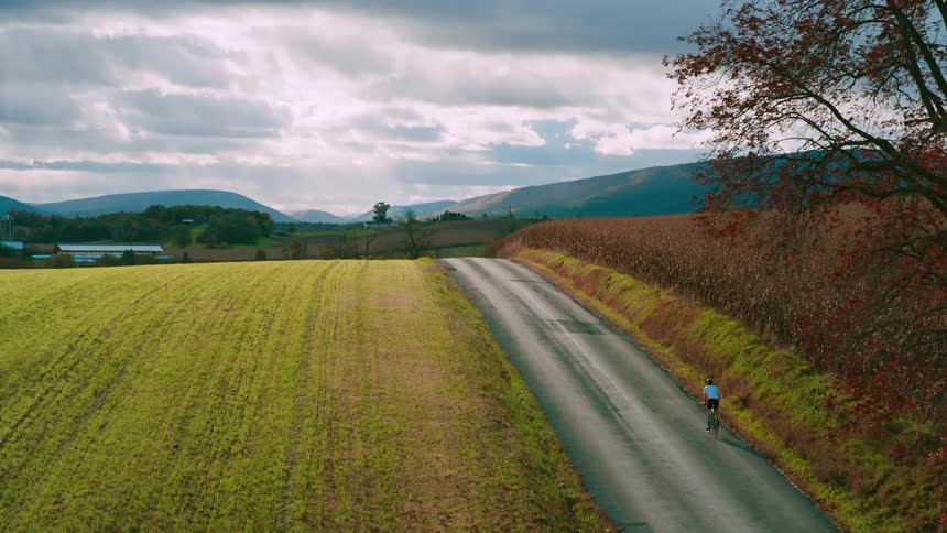 A cyclist rides uphill on a narrow country road between a green field and distant mountains under a cloudy sky
