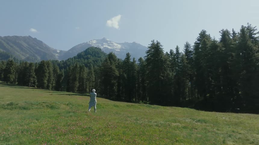 Person with white turban and long shirt walks across a green grass field with trees behind and mountains on the horizon