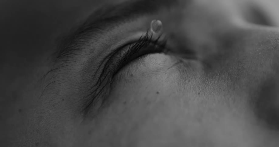A drop of water falling into the eye of a person who has their eyes closed and everything is in black and white tones