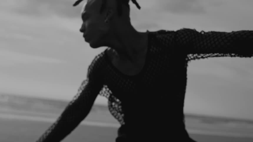 A person wearing a netted longsleeve top dances on a beach with the ocean and sky in the background