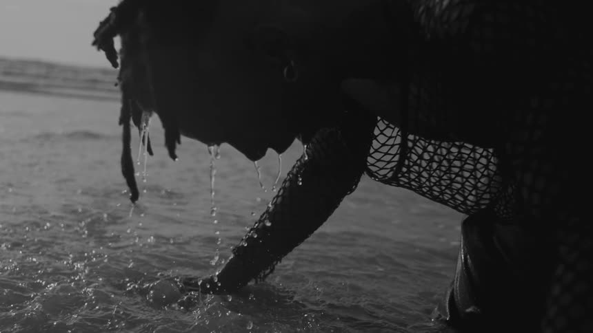 A man with wet hair and a mesh sleeve is leaning over and touching the surface of the water with droplets falling from the hair and face