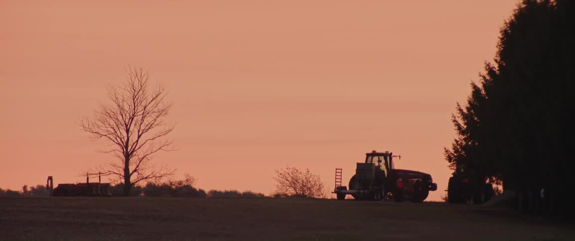 A tractor is silhouetted against a peachcolored sky at dusk standing on a grassy field next to bare trees