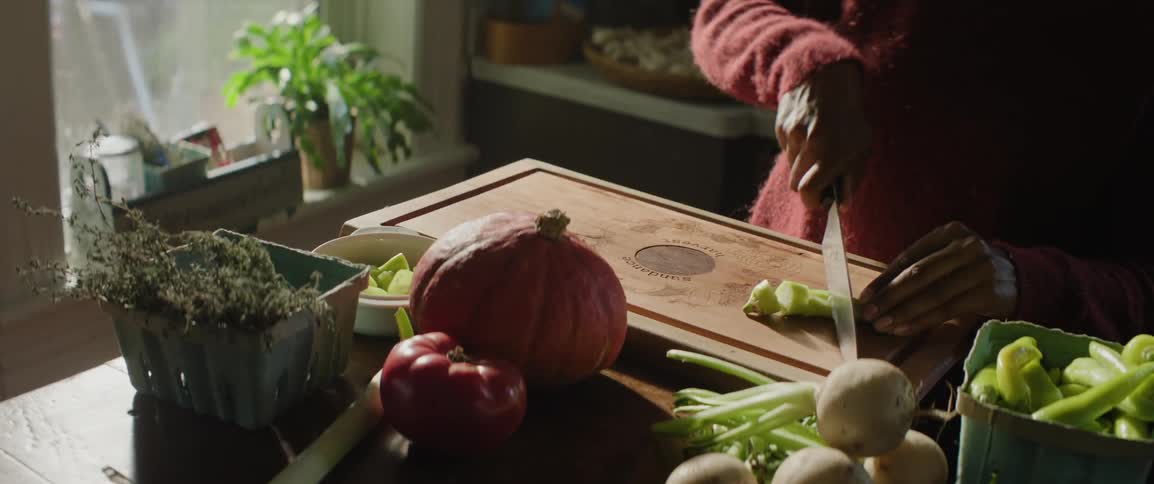A person in a red sweater stands at a cutting board in a sunlit kitchen chopping a pepper alongside other fresh produce near a window with potted plants