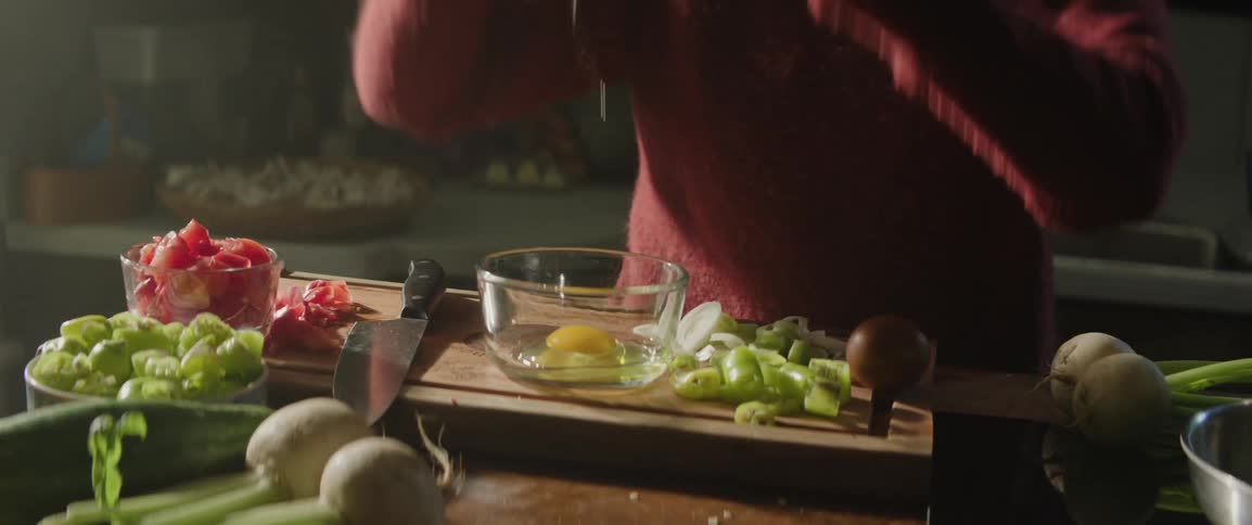 Woman in a red sweater cracks brown eggs into a glass bowl on a cutting board with a knife with freshly chopepd onions and sliced vegetables in a kitchen