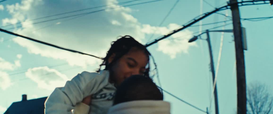 A father in a white hoodie lifts his longhaired daughter into the air with the sun shining brightly behind them