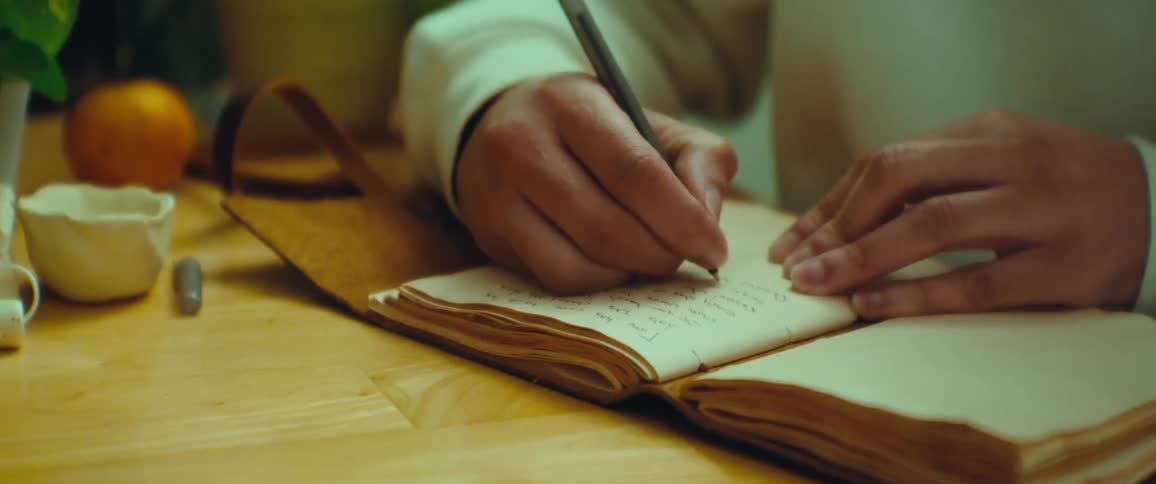 A person writes in an old worn notebook with a pen on a wooden table