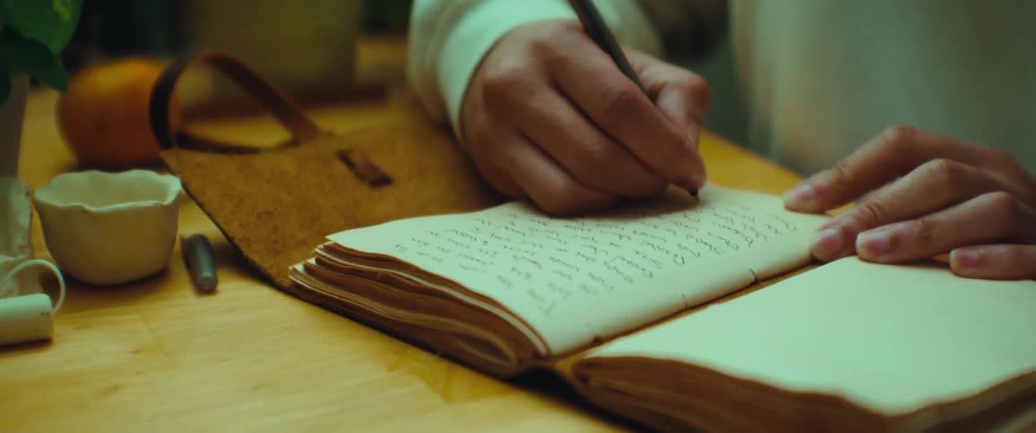 A person writes in an old worn notebook with a pen on a wooden table