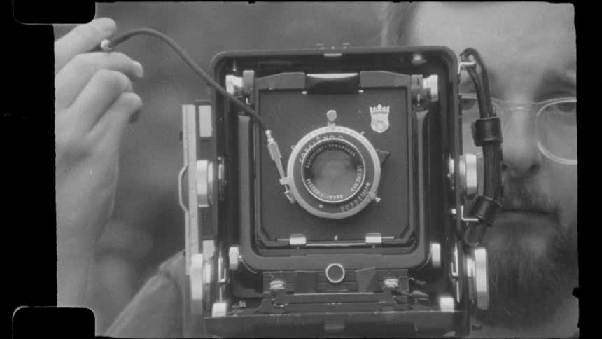 The face of a bearded man wearing glasses who is taking a picture using an old camera