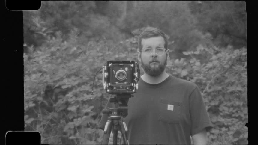 Bearded man with glasses poses next to an analog camera mounted on a tripod amid vegetation