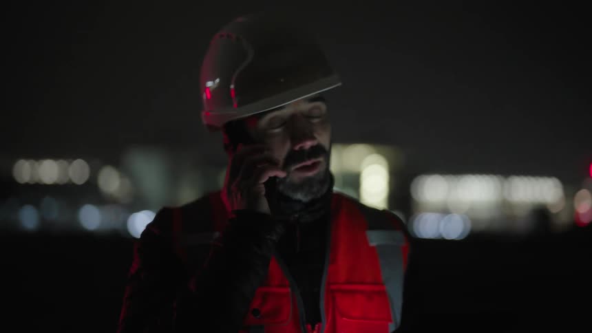 A person wearing a hard hat and a reflective vest uses a smartphone at night with an illuminated cityscape in the background