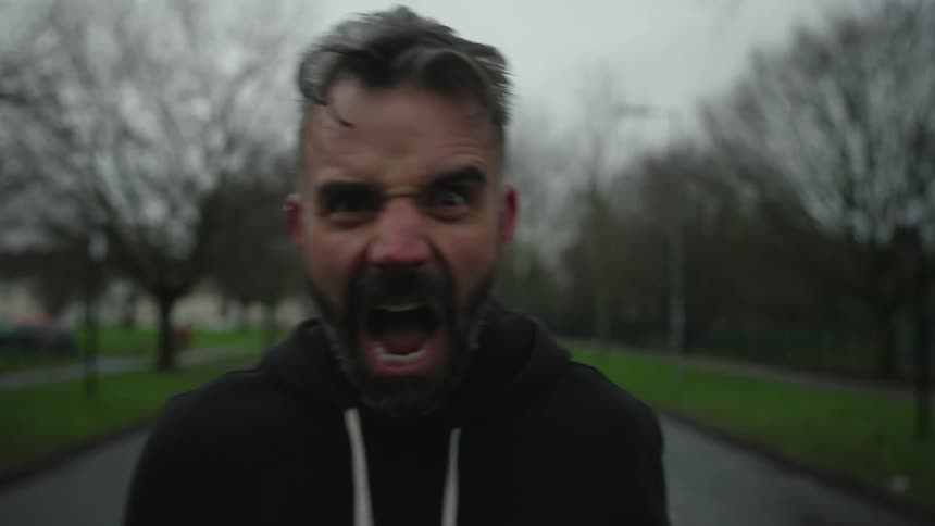 A bearded man stands on a path in a park screaming at the camera