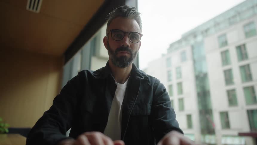 A bearded man in glasses typing on a laptop near an office window