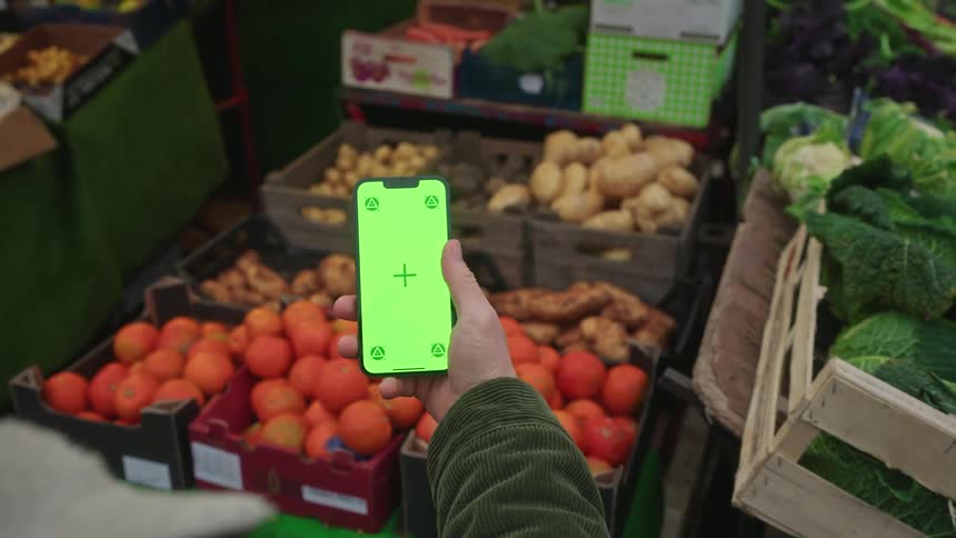 A person holds a smartphone with a green screen display in front of producefilled crates at a market