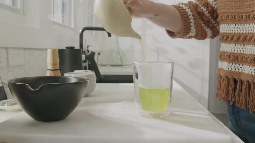 A person pours a tea from a teapot into a clear glass on a white countertop in the kitchen