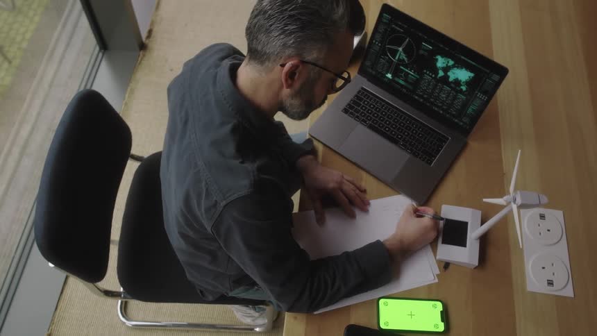 Man writes in a notebook next to a laptop with data analysis graphs a small wind turbine and a smartphone