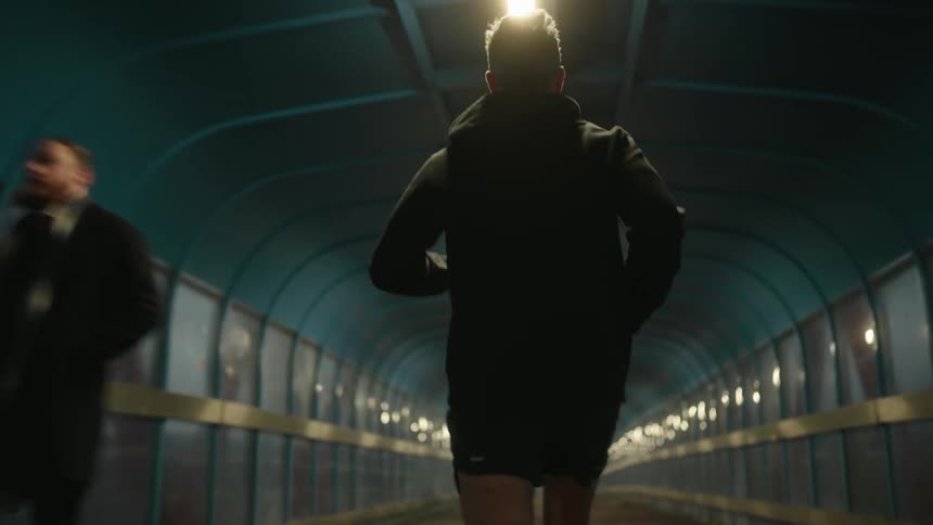 Man in a black hoodie runs through a lit tunnel while two people walk in the opposite direction