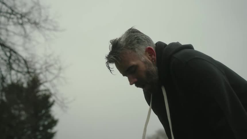 Man in a black hoodie bends over breathlessly outdoors under a cloudy sky and starts running again