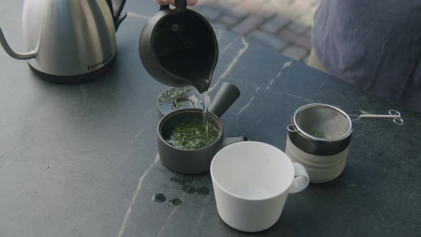 Person pours tea from a black teapot over tea leaves in a container with a strainer and white cup next to it
