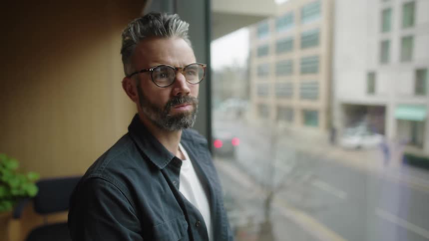 Closeup of a bearded man with glasses looking out the office window at the city