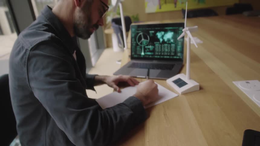 Man sitting at an office desk with an open laptop and writing on a sheet of paper