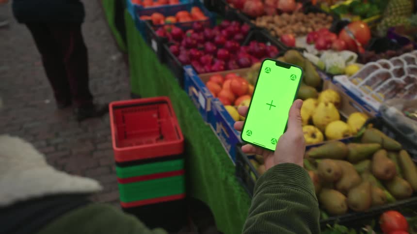 Person holds a smartphone with a green screen in front of a fruit stand at an outdoor market