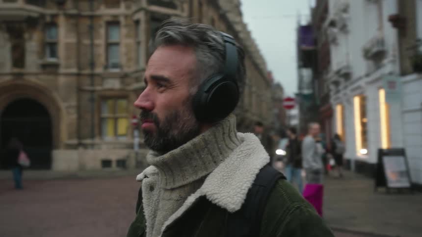 A bearded man wearing headphones and a wool coat walks down a busy street at dusk