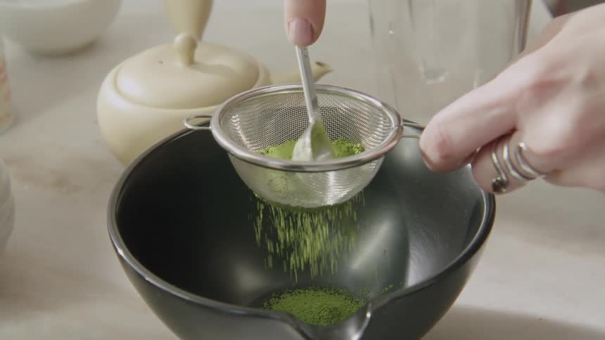A woman sifts matcha powder through a strainer into a black tea bowl with a white teapot visible in an indoor tea preparation setting