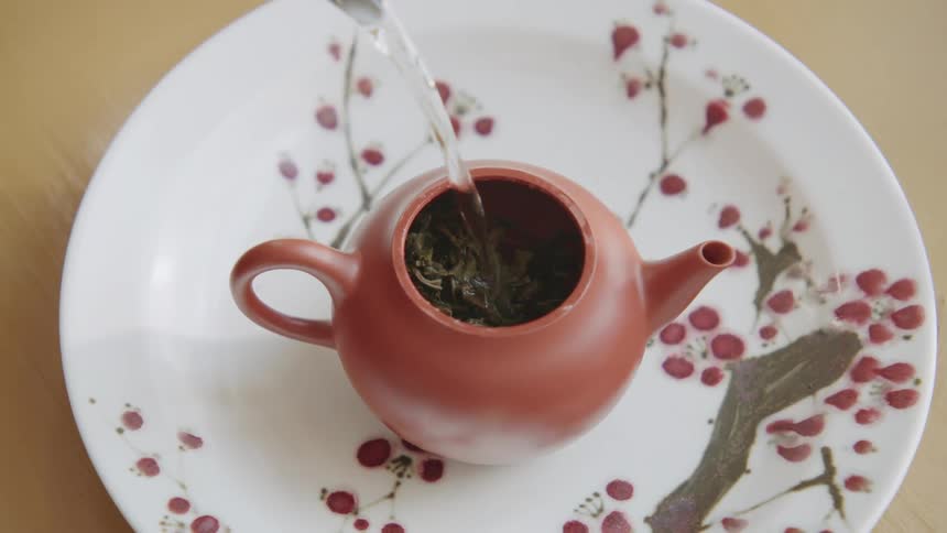 A closeup of a person pouring hot water into a small teapot filled with tea leaves