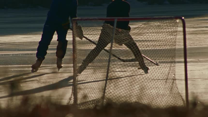 Two men playing hockey on an ice surface and one of them is attacking while the other is defending in goal