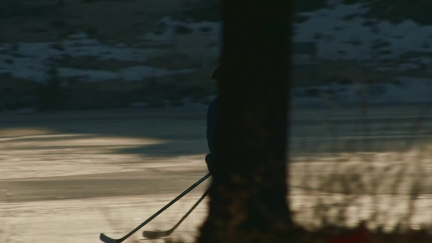 Two silhouetted hockey players perform drills powerfully skating across a glassy frozen lake towards a red hockey goal surrounded by snowdusted trees all bathed in soft morning sunlight