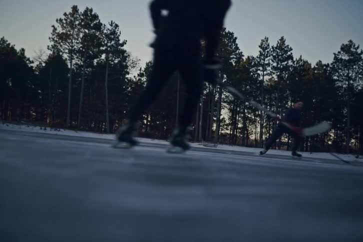 Two hockey players skate on an outdoor rink surrounded by tall pine trees