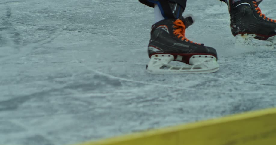 A closeup shows a person wearing ice skates with orange laces sharply turning on an outdoor rink and kicking up ice near a yellow guardrail