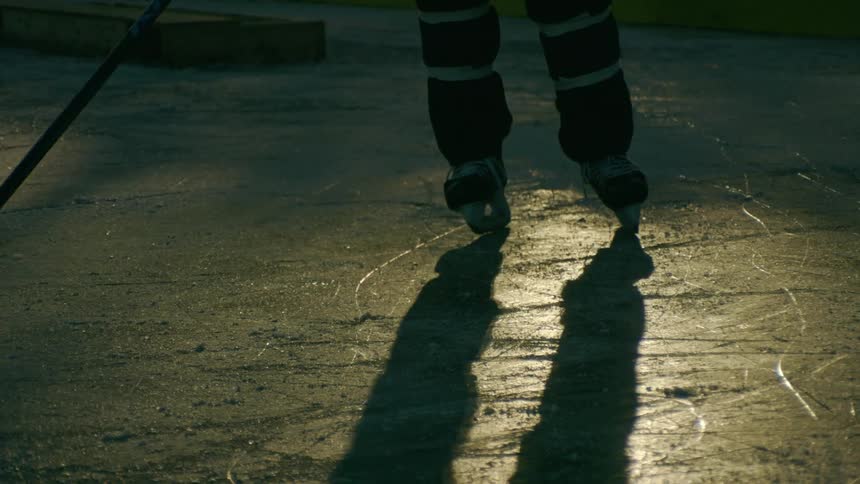 A hockey players legs and skates on the ice casting long shadows with a stick and puck visible