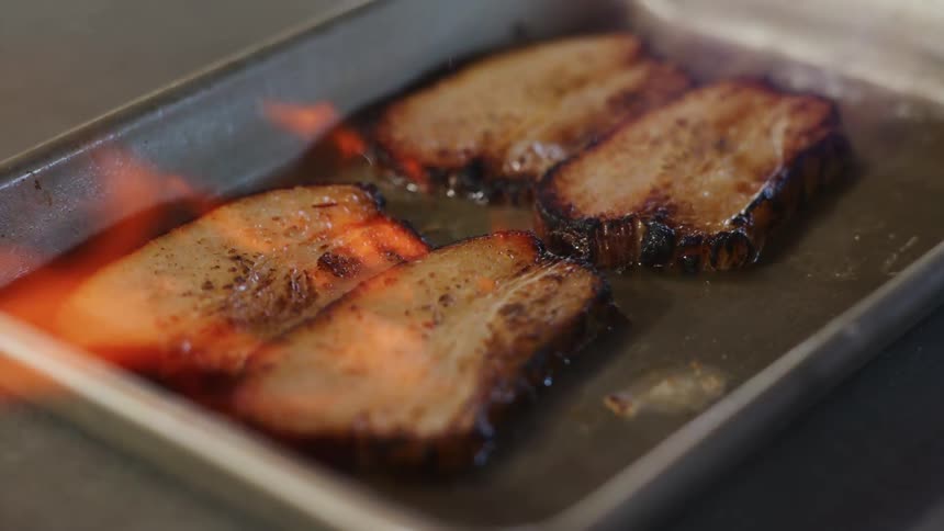Slices of pork are being fireblasted in a silver pan with flames licking the sides creating char marks as the food cooks