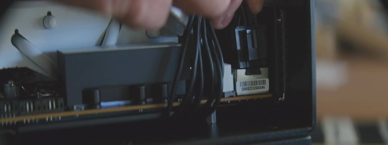 A hand is shown plugging a black cable into a port on a computer component inside a black casing