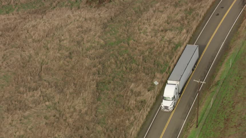 An aerial view shows a white semitruck driving on a narrow road through fields in the countryside