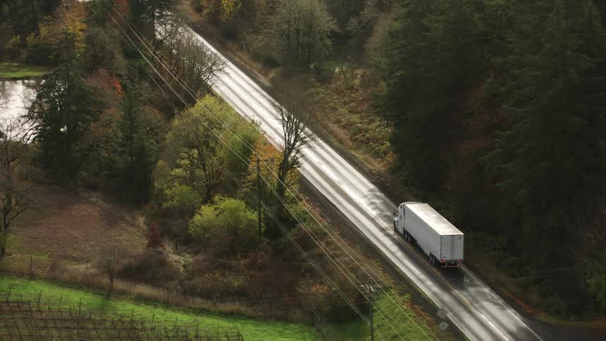 A white semitruck drives along a winding rural highway bordered by farmland trees and power lines