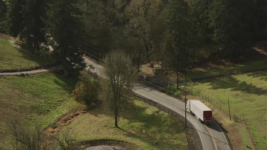 An aerial view shows a white semitruck driving on a narrow road through a wooded area