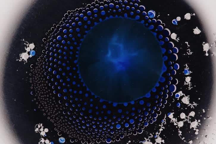 A dark circular center contract inwards surrounded by a ring of white droplets and a lighter outer edge creating a dynamic abstract pattern