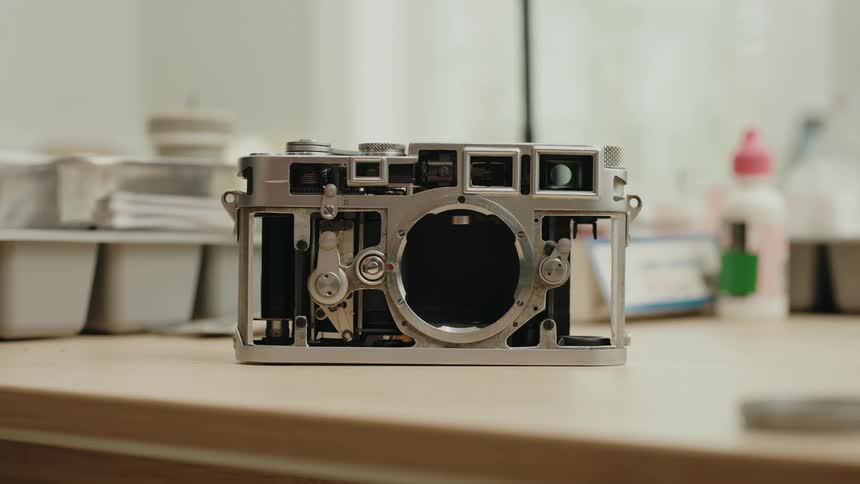 A disassembled silver camera body revealing its internal mechanisms sits on a welllit workshop table