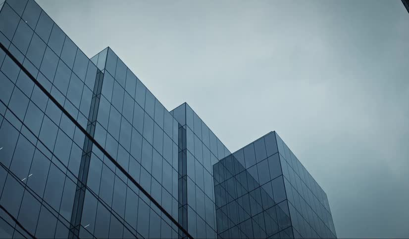 An upward shot shows the angular facade of a modern building with reflective glass panels against a gray sky