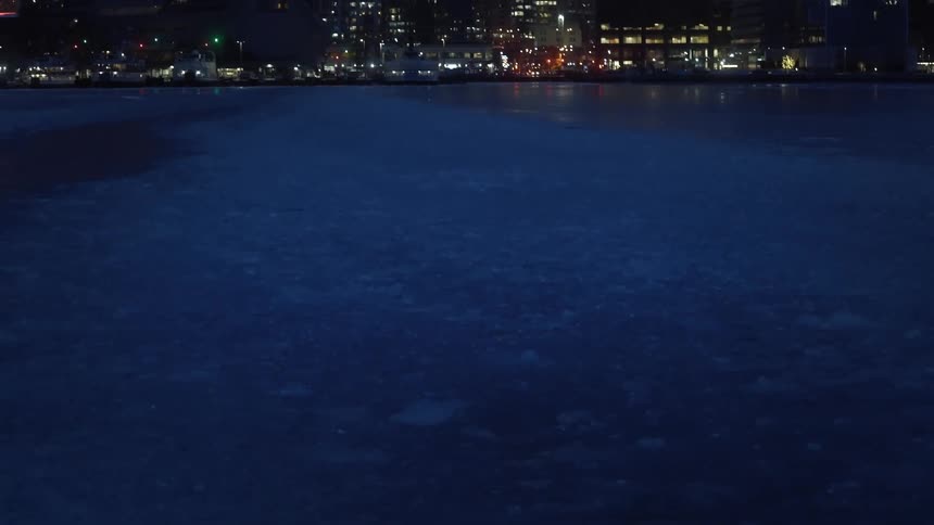 Partially frozen lake reflects the lights of illuminated skyscrapers at night in the city