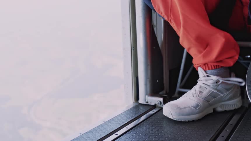 A person wearing orange pants and white sneakers is sitting at the open door of an aircraft with one foot resting on the metal floor